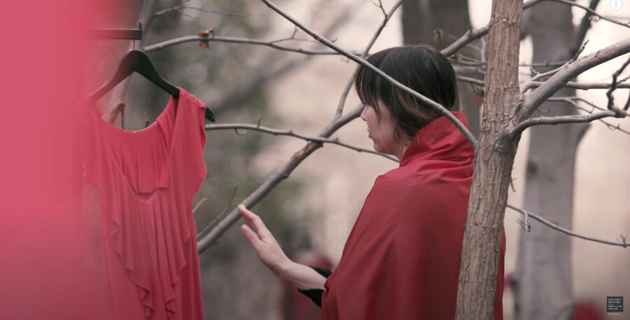 Jaime Black a Métis artist stands with a red dress draped around her as they move to touch another that hands from a tree. Another red dress can be seen blurry in both the foreground, and many more in the background.