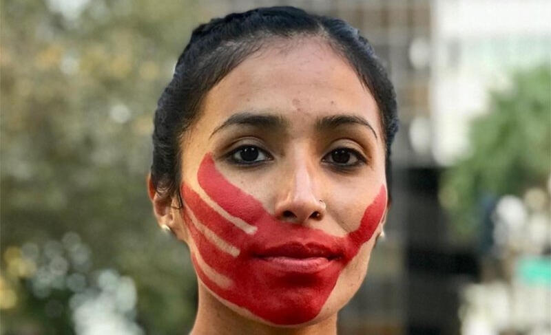 Jordan Marie Brings Three White Horses Daniel, an Indigenous athlete faces the camera with a stoic face as a red handprint of paint lays across their lips and cheeks as if being silenced by the paint.